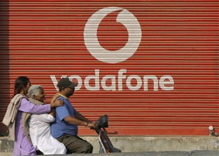 A man and two passengers ride on a scooter past a shop displaying the Vodafone logo on its shutter in Jammu