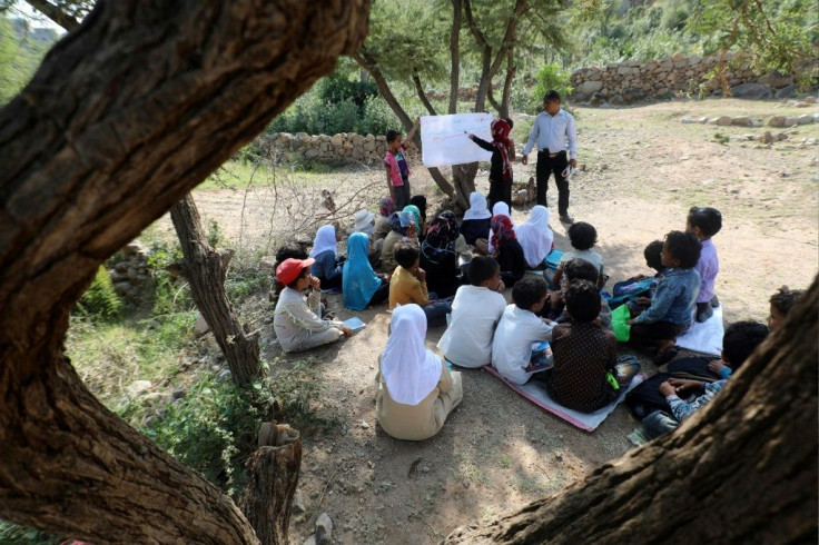 Twenty Yemeni pupils listen to their teacher with textbooks balanced in their laps as they sit under trees in their southern village, where the school building remains unfinished