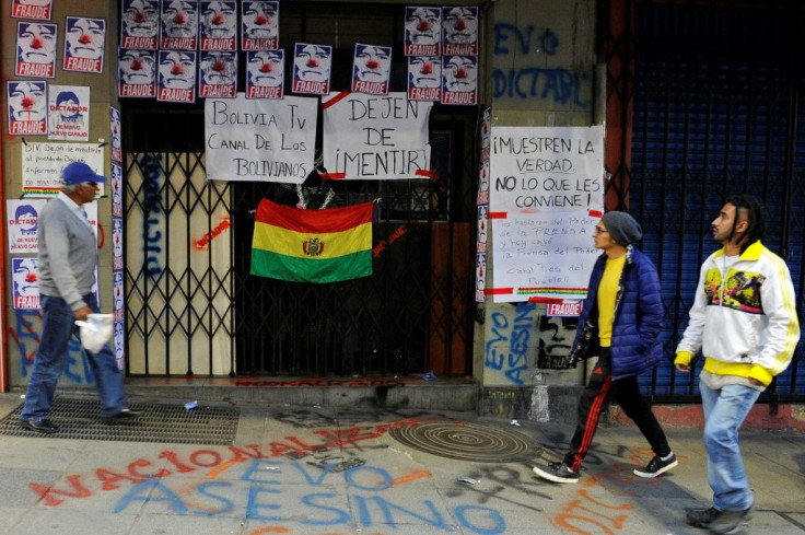 People walk past the closed entrance of state TV channel BoliviaTV, plastered with pictures depicting President Evo Morales as a clown and calling him a "fraud" on November 9, 2019, in La Paz