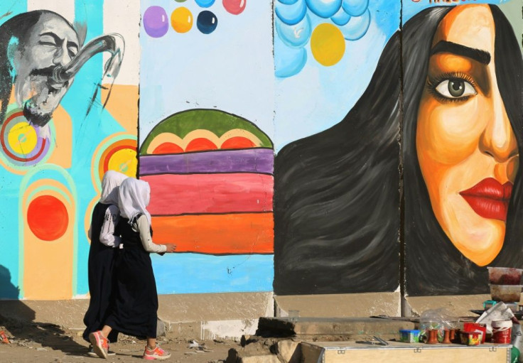 Iraqi women walk past a mural inspired by ongoing anti-government protests in Tahrir square in Baghdad while political leaders are struggling to choose a new prime minister