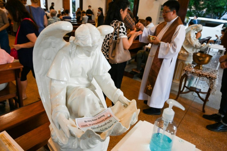 A priest hands a holy communion in front of a statue of an angel with a sign saying "no holy water during COVID-19 precautionary period" and a bottle of hand sanitiser at a church in Bangkok