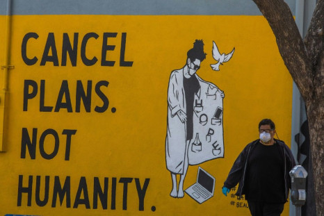 A man wearing gloves and a face mask walks by a mural reading "Cancel Plans Not Humanity" in Los Angeles, California