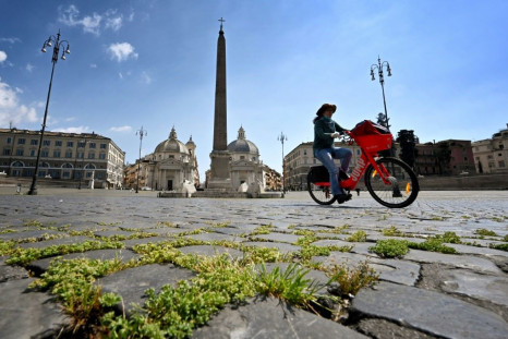 Grass grows between cobblestones as a woman rides a bicycle across Piazza del Popolo on April 24, 2020 in Rome, during the country's lockdown aimed at stopping the spread of the COVID-19 pandemic, caused by the novel coronavirus