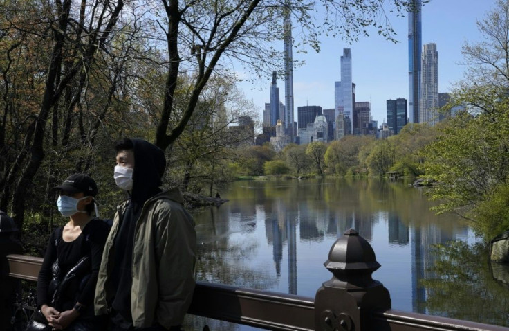 The Midtown Manhattan skyline is reflected in the water as people in masks walk along the lake in Central Park