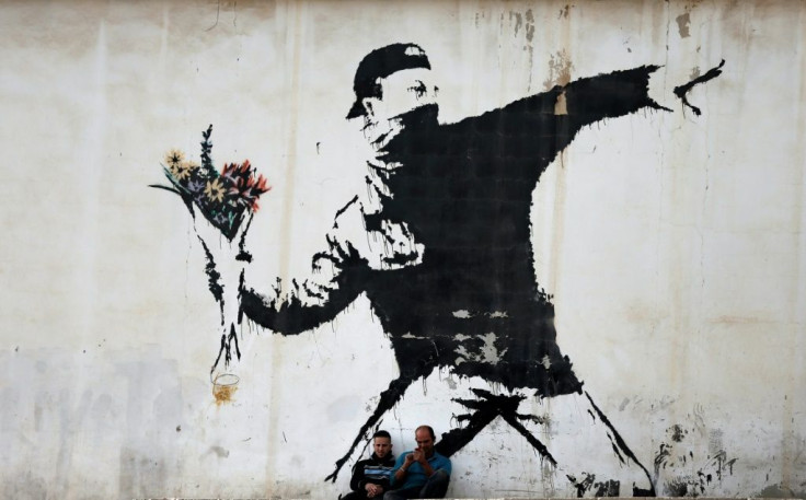 Two men sit in front of Banksy's 'Flower Thrower' mural painted on a wall of a petrol station in the West Bank town of Bethlehem