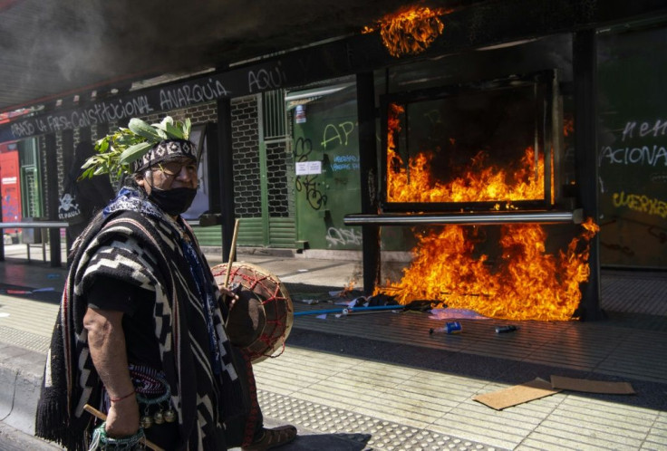 A Mapuche indigenous man plays a tambourine as he marches past an advertisement set on fire by demonstrators who clashed with riot police during a protest in Santiago