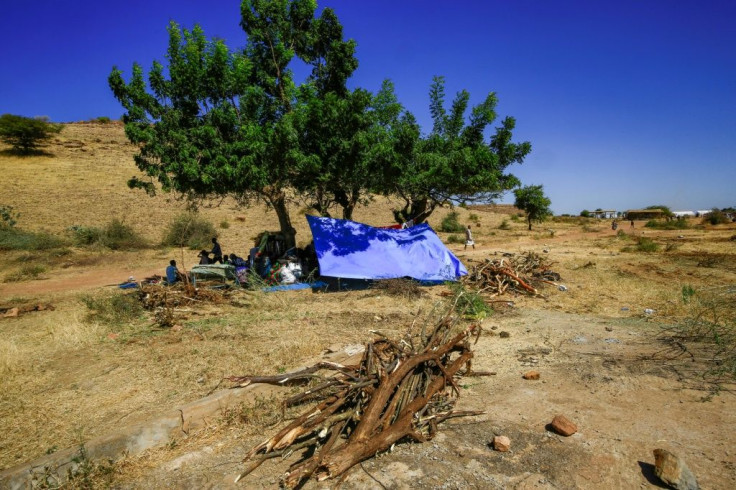 People sit in the shade of a tree next to mattresses, beds, and belongings at the Um Raquba camp in Sudan's eastern Gedaref province, where thousands of refugees have fled to from Ethiopia's Tigray conflict