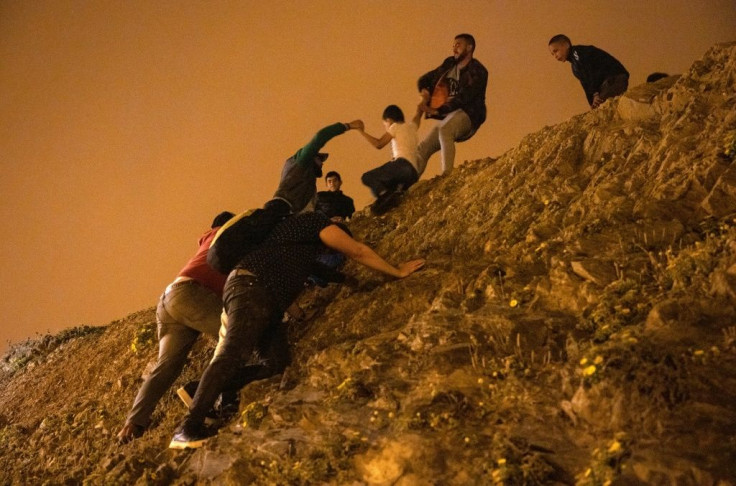 Moroccan migrants climb a rocky cliffside as a policeman watches in the northern town of Fnideq as they attempt to cross the border from Morocco to Spain's North African enclave of Ceuta on May 18, 2021.At least 5,000 migrants, an unprecedented influx at