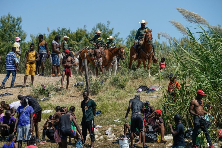 United States Border Patrol agents on horseback look on as Haitian migrants sit  on the banks of the Rio Grande near Del Rio, Texas.