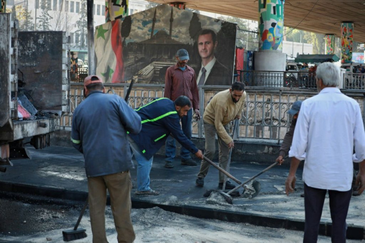 Municipal workers clean up after the bombing with a roadside portrait of Syrian President Bashar al-Assad looming in the background
