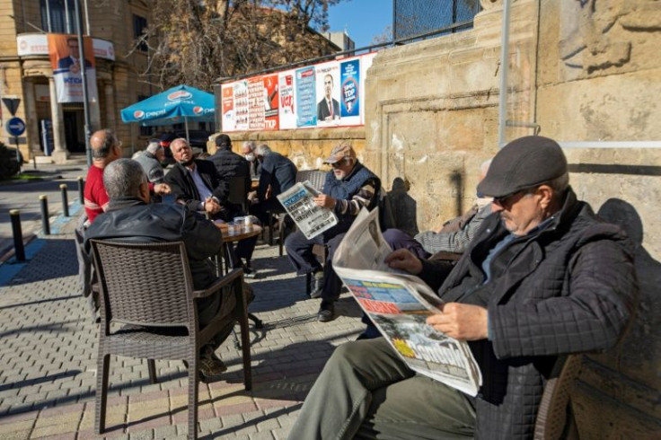 People sit at a cafe next to election posters in the northern part of Cyprus's divided capital Nicosia, in the self-declared Turkish Republic of Northern Cyprus, ahead of the general election dominated by economic issues