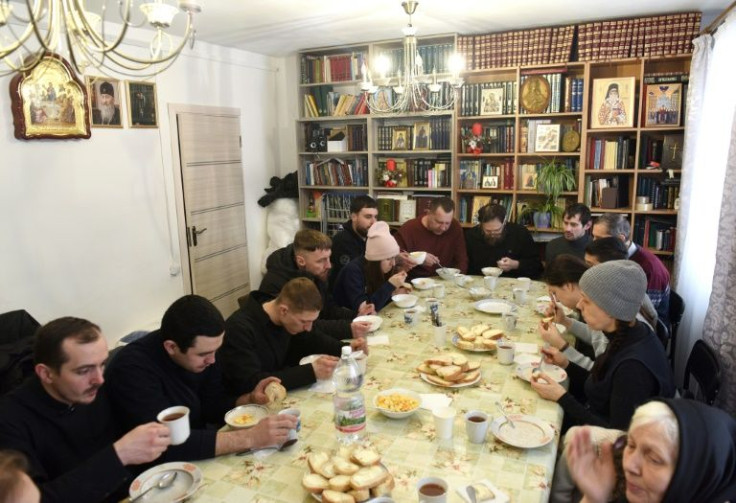 Refugees take their breakfast in a room filled with books and icons