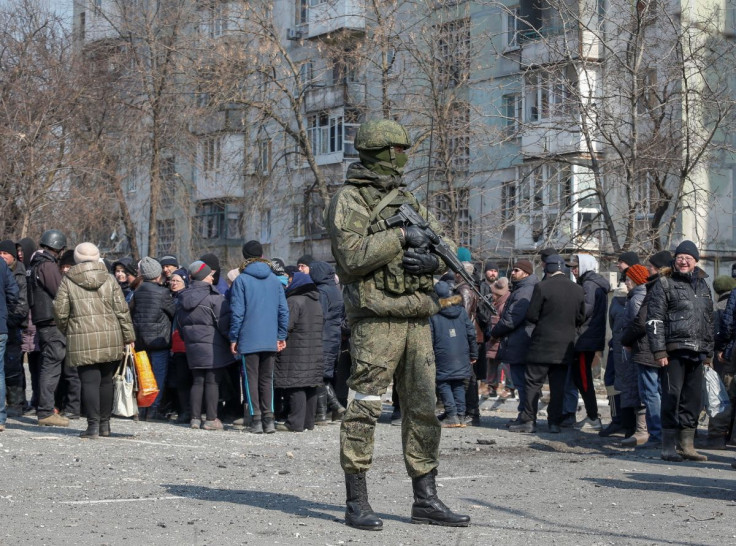 A Russian Army soldier stands next to local residents as they line up to deliver humanitarian aid during the Ukraine-Russian conflict in the besieged southern Ukrainian port of Mariupol, March 23, 2022.  
