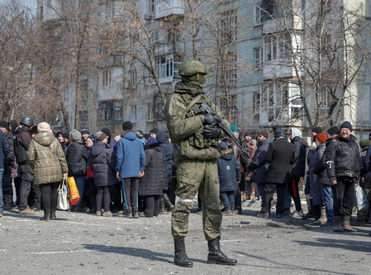 A Russian army soldier stands next to local residents who queue for humanitarian aid delivered during Ukraine-Russia conflict, in the besieged southern port of Mariupol, Ukraine March 23, 2022.  