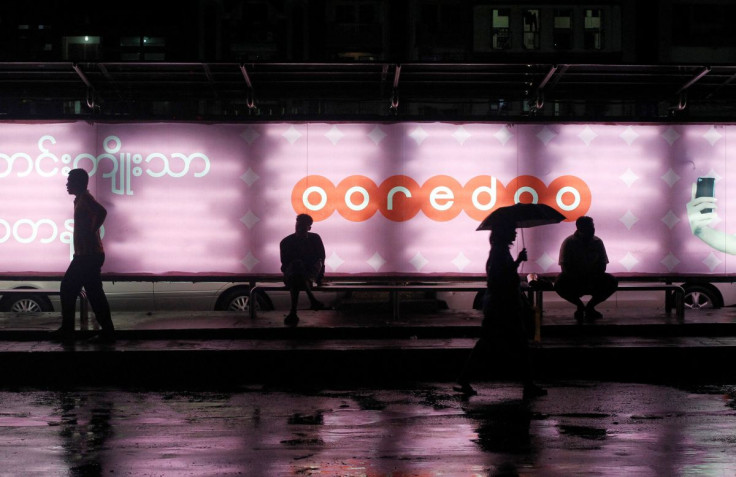 People sit at a bus station with an Ooredoo advertisement as they wait for a bus in Yangon, August 1, 2014.