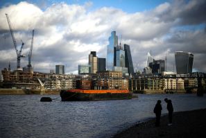 People stand on the bank of the River Thames with the City of London financial district in the background, in London