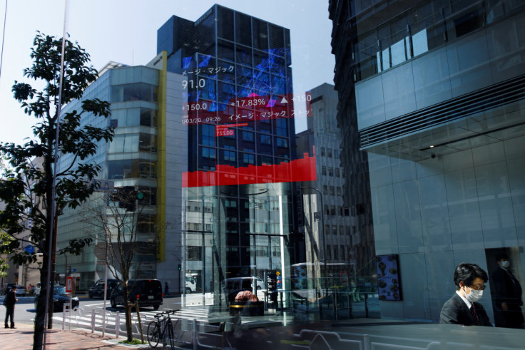 A man uses a laptop, under an electronic board showing stock visualizations, inside a brokerage building, in Tokyo