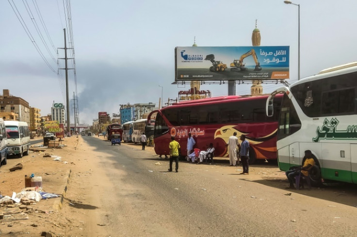 Smoke billows in the distance as people wait next to passenger buses in Sudan's capital Khartoum