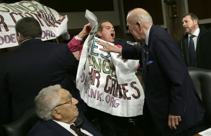 Protesters demand the arrest of former secretary of state Henry Kissinger as he sits to testify in 2015 at a Senate hearing but the banner is deflected by another former top US diplomat, George Shultz