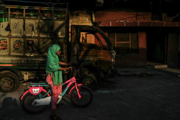 A girl rides a bicycle past a charred vehicle belonging to a local Muslim family burnt during the anti-Muslim riots in February