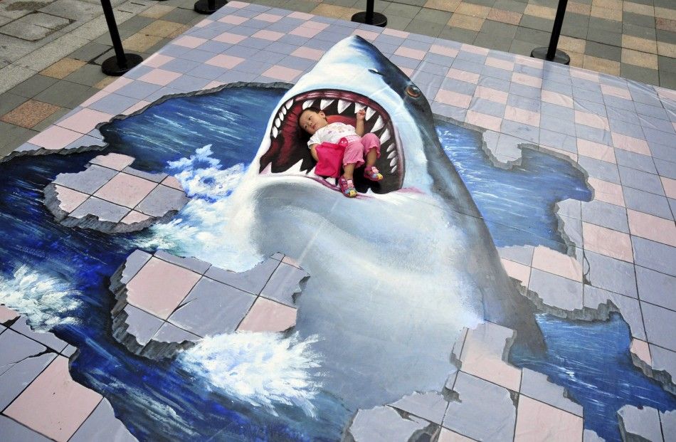 A child lies on a piece of 3D pavement artwork featuring a shark as she poses for a photograph for her family inside a shopping mall in Fuzhou