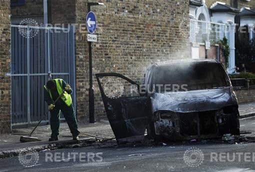 A street cleaner sweeps up around a smouldering van set alight during riots in Hackney in London